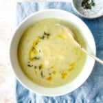 celery and leek soup in a white bowl with a spoon in it on a blue cloth next to a bowl of herbs.