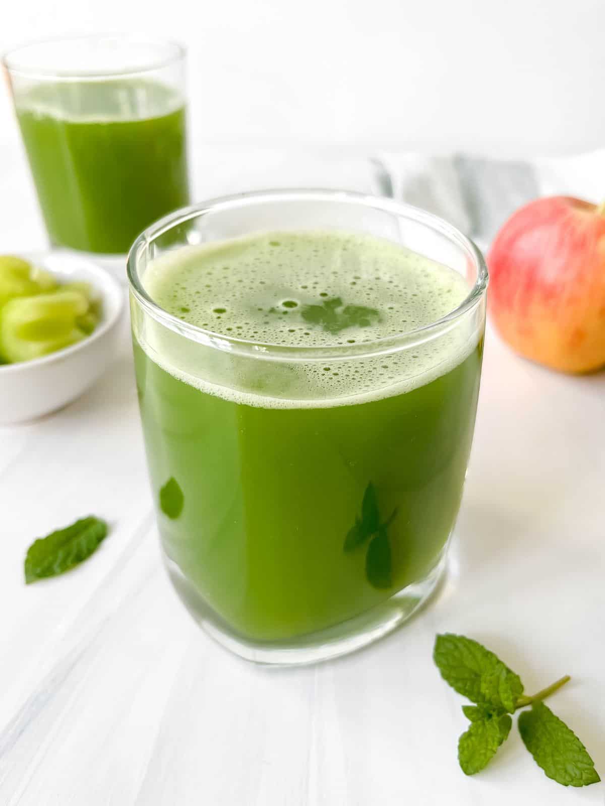 celery cucumber juice in two glasses next to mint leaves and a red apple.