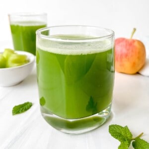 celery cucumber juice in two glasses with a red apple in the background.
