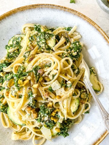 creamy kale pasta in a light grey bowl with a spoon next to a small bowl of pistachios.