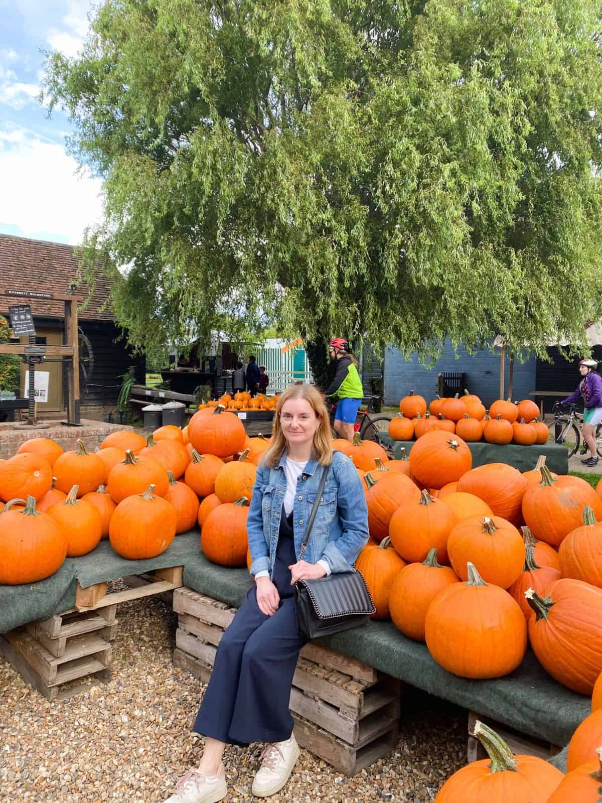 woman wearing a denim jacket sitting down with lots of pumpkins behind her.