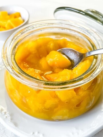 mango compote in a glass jar with a spoon in it on a white plate.