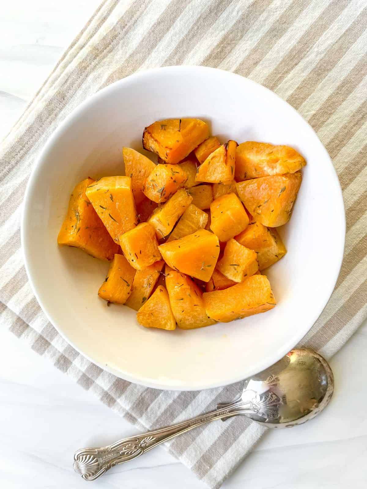 butternut squash cubes in a white bowl on a beige and white striped cloth next to a spoon.