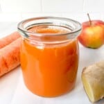 carrot ginger turmeric juice in a glass next to carrots, ginger and a red apple.