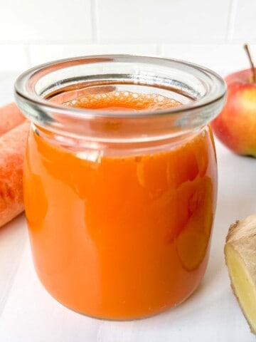 carrot ginger turmeric juice in a glass next to carrots, ginger and a red apple.