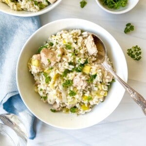chicken and leek risotto in a white bowl with a spoon in it next to a blue cloth and glass of water.