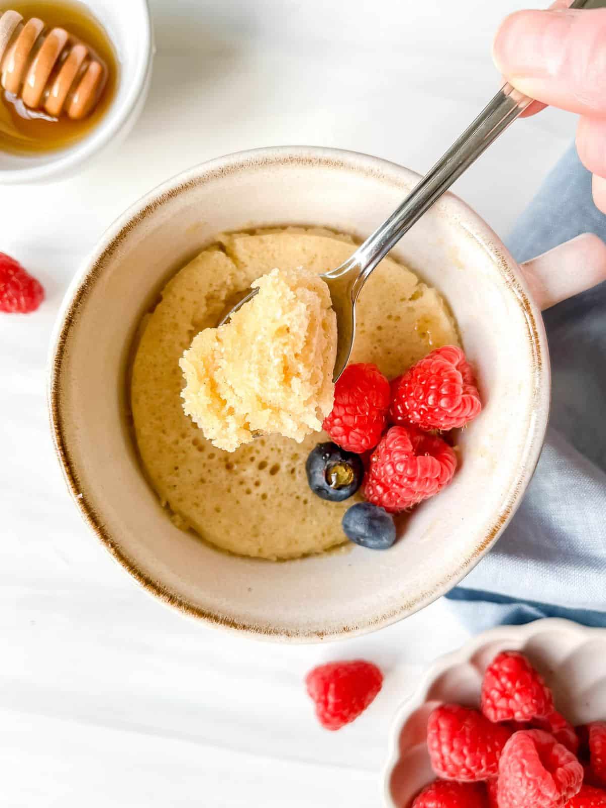 honey mug cake in a light brown mug with a spoonful above it next to a bowl of raspberries.