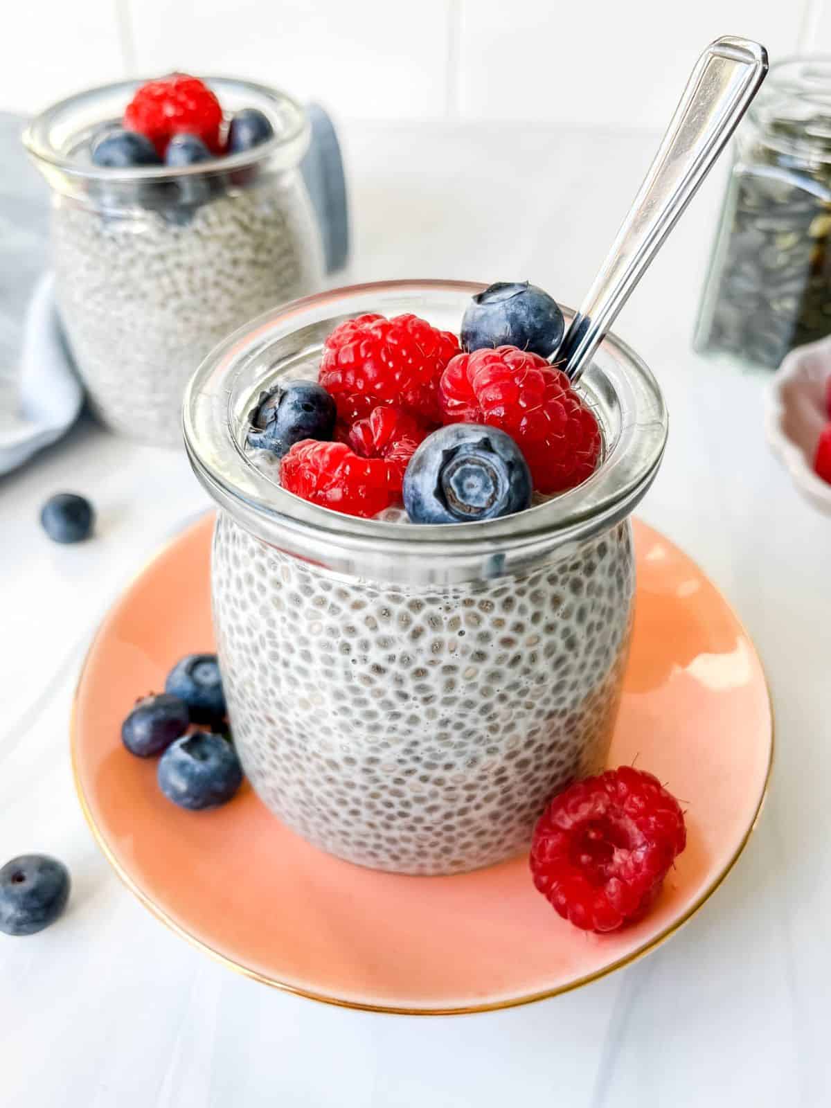 two glass jars of oat milk chia pudding with one on a pink plate topped with berries.