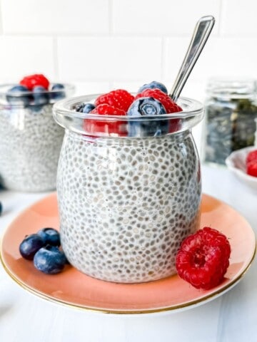 oat milk chia pudding in two glass jars with one on a pink plate topped with berries and a jar of pepitas in the background.