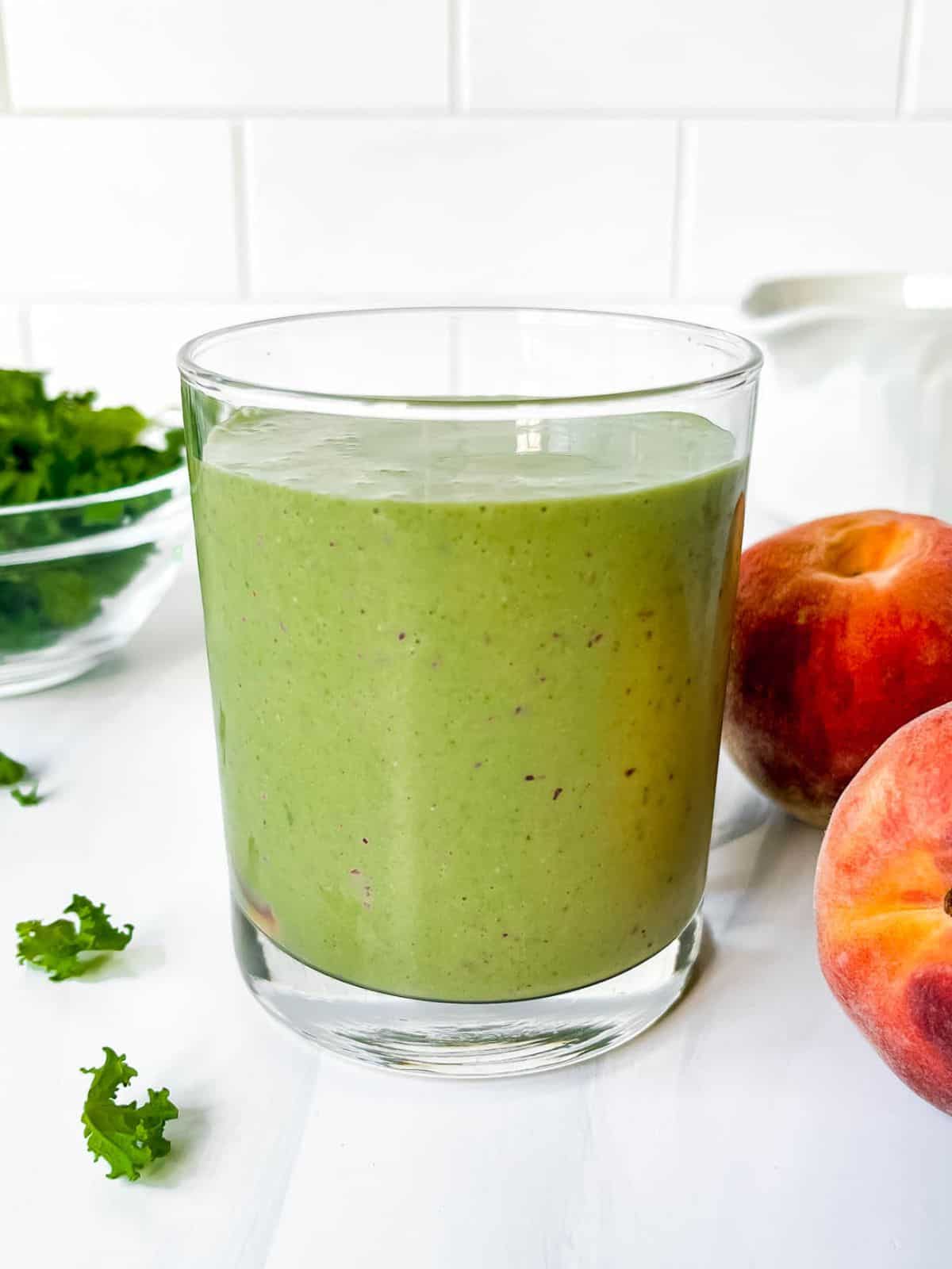 peach kale smoothie in a glass next to peaches and a bowl of kale leaves.