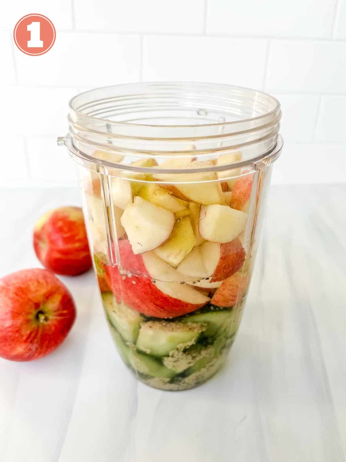 apples, cucumber, seeds and water in a blender cup next to red apples labelled number one.