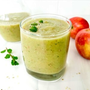 apple cucumber smoothie in a glass and jug next to red apples and mint leaves.
