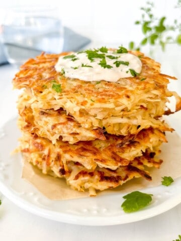 stack of grated potato fritters on a white plate with fresh herbs and glass of water in the background.