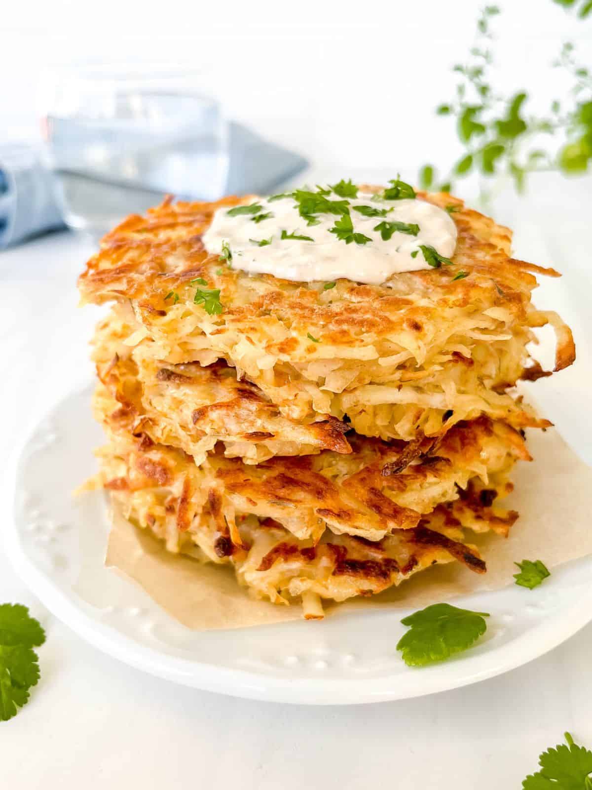 grated potato fritters in a stack on a white plate next to herbs and a glass of water.
