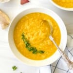 parsnip and ginger soup in a white bowl garnished with fresh herbs with a spoon in it next to ginger and garlic.