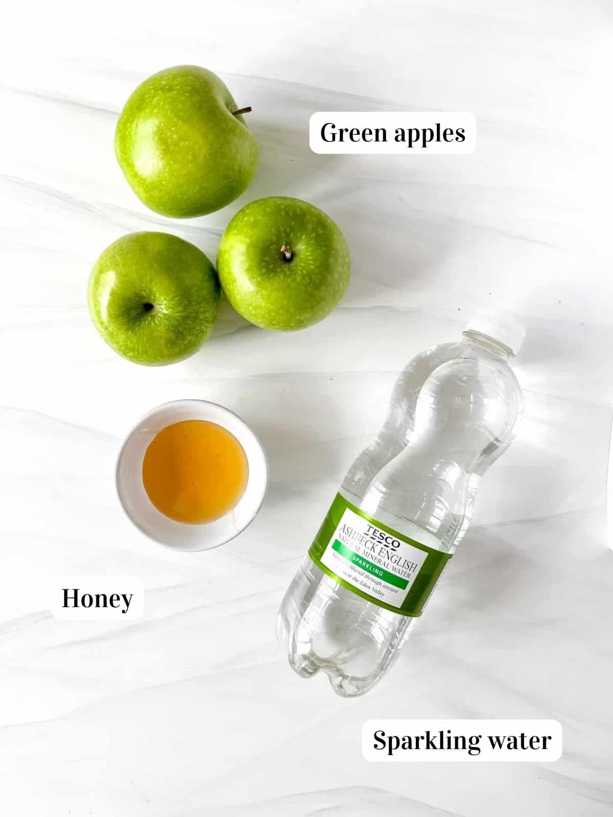 individually labelled green apples, bottle of sparkling water and small white bowl of honey.