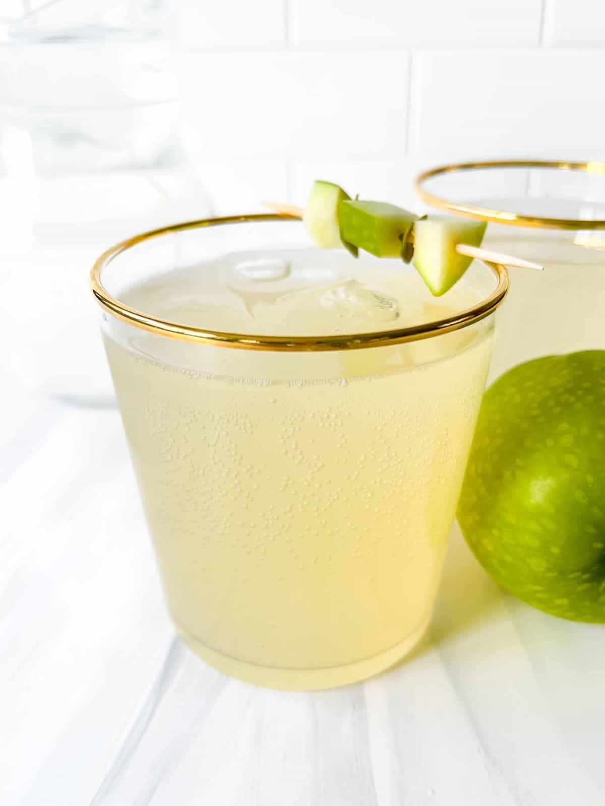 green apple mocktail in two glasses with gold rims next to a Granny smith apple.