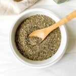 Italian seasoning in a white bowl with a wooden spoon in it next to a beige cloth.