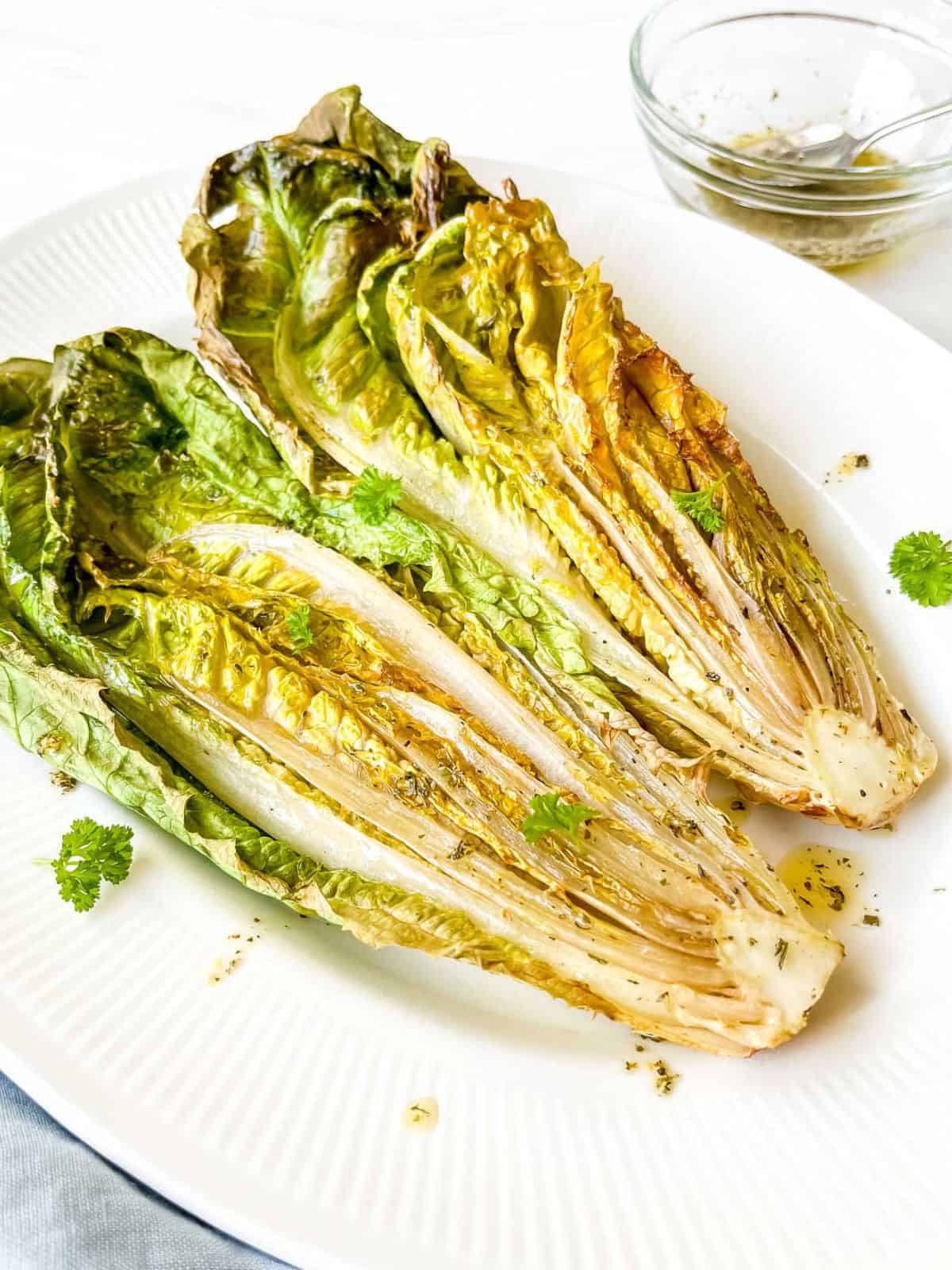 two halves of romaine lettuce on a white plate with a glass bowl of oil dressing in the background.