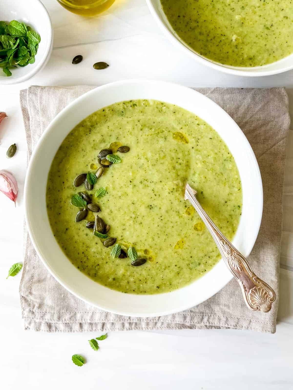 broccoli zucchini soup in two white bowls on a beige cloth next to a bowl of fresh mint and garlic cloves.
