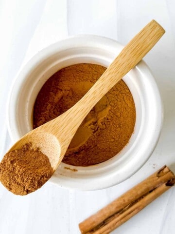 wooden spoon of cinnamon on top of a white bowl of cinnamon next to a cinnamon stick/