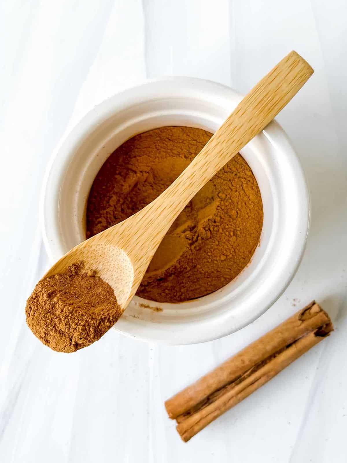 a spoonful of cinnamon on top of a bowl of ground cinnamon next to a cinnamon stick.