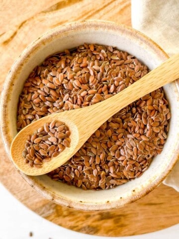 bowl of flaxseeds in a small cream bowl with a spoonful of seeds on top of it on a wooden board and beige cloth.