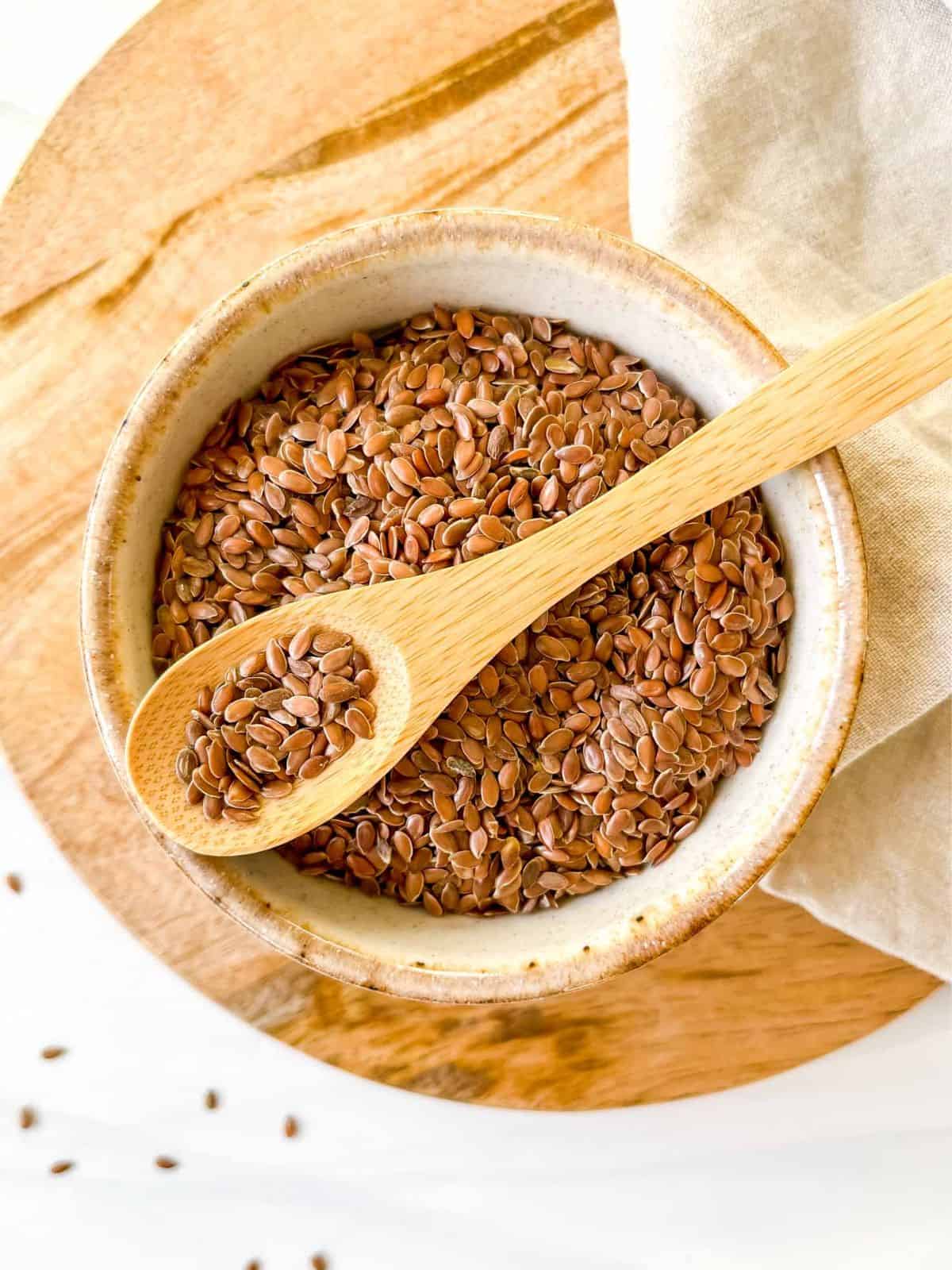 brown flaxseeds in a cream bowl with a spoonful of flaxseeds on top of it on a wooden board and beige cloth.