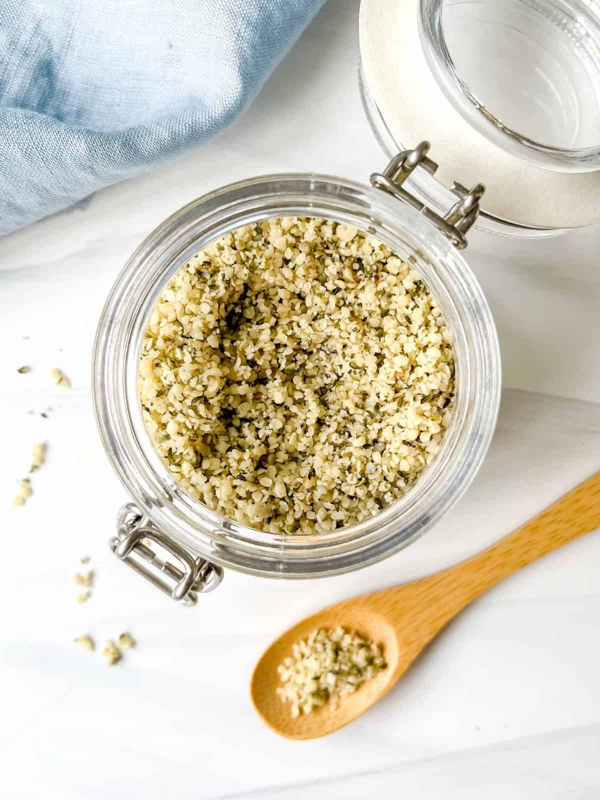 hemp seeds in a glass jar next to a blue cloth and spoonful of seeds.
