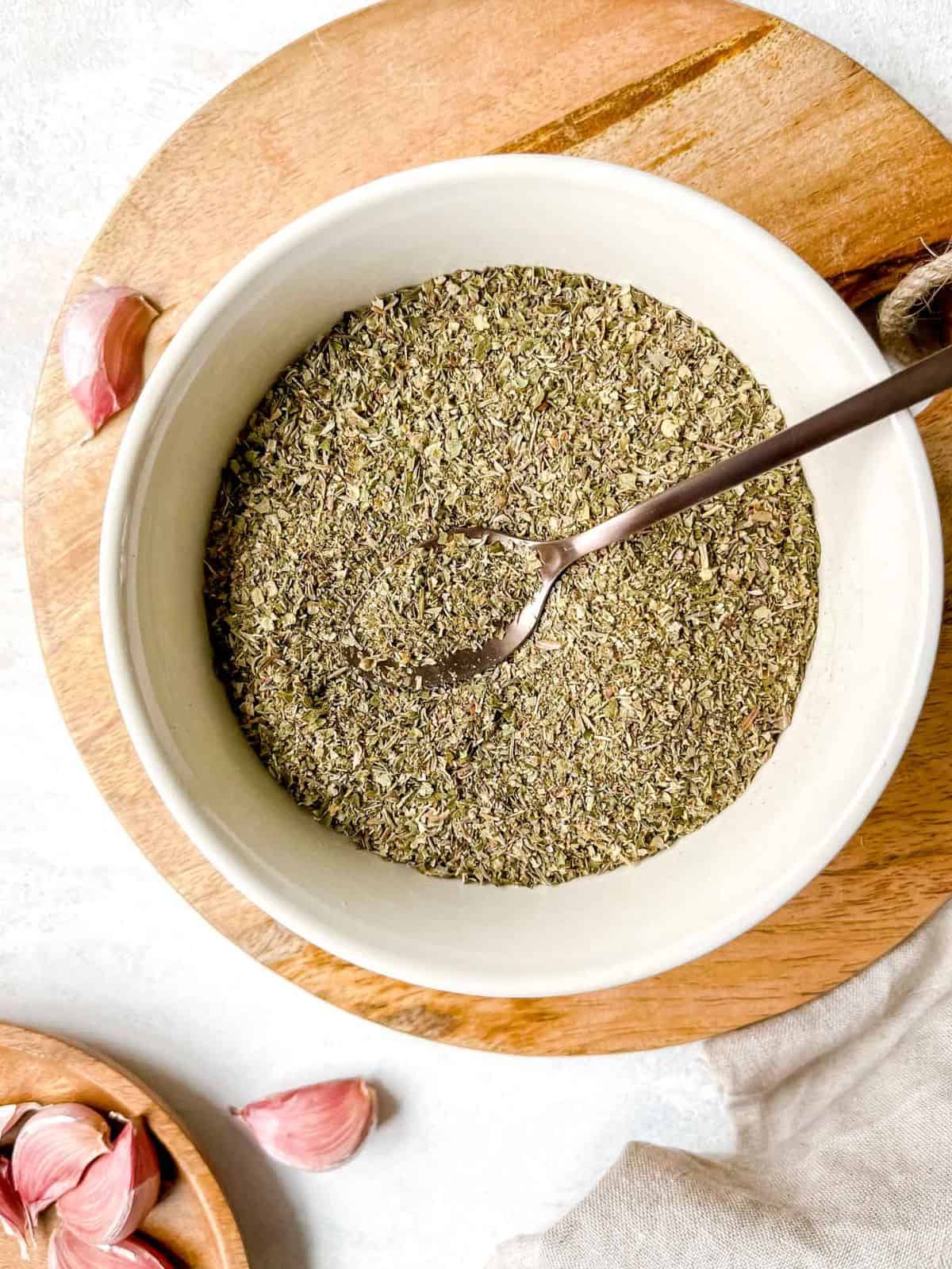 herbes de Provence in a cream bowl with a spoon in it on a wooden board next to garlic cloves.