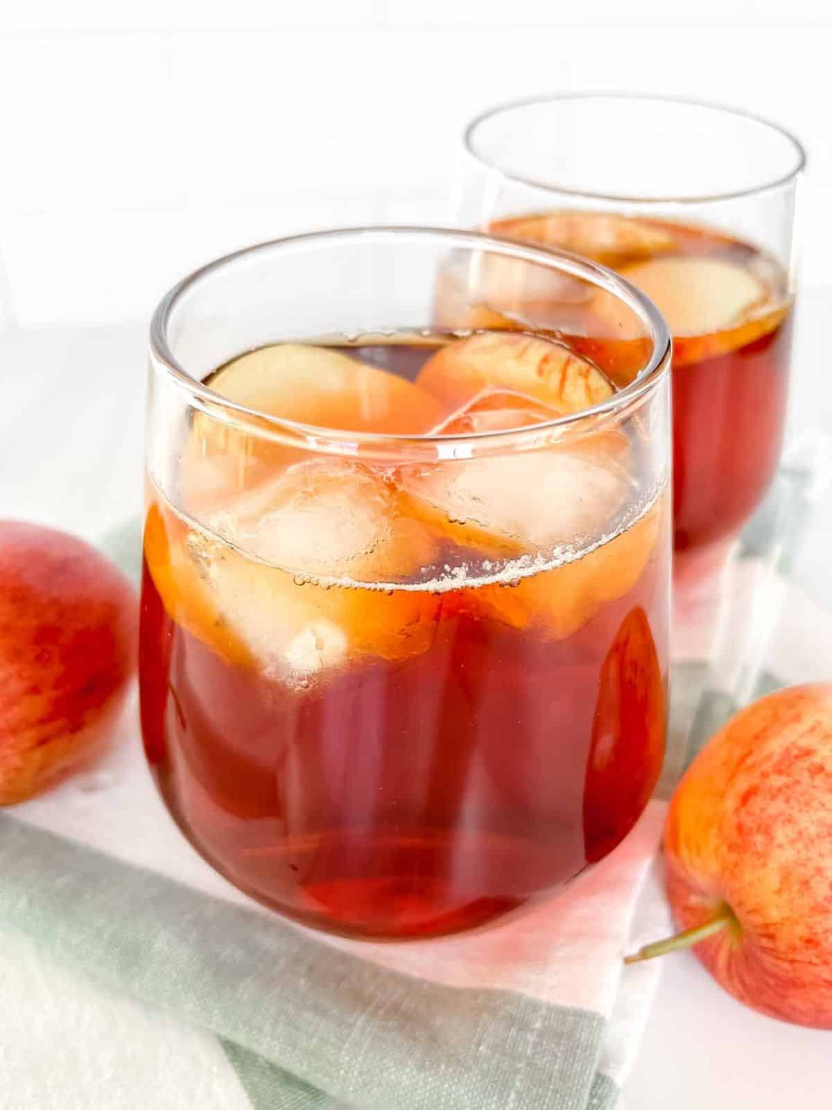 iced rooibos tea in two glasses on a white and green striped cloth next to red apples.