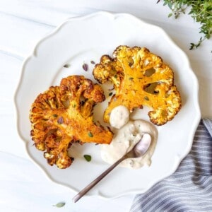 two cauliflower steaks on a white plate with tahini and a spoon on a blue cloth.