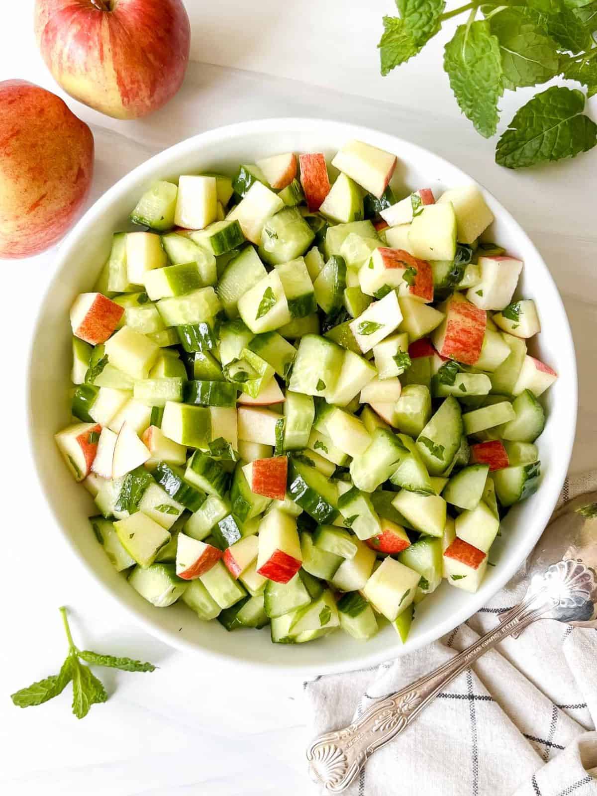 apple and cucumber salad in a white bowl next to a spoon, red apple and mint leaves.