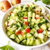 apple and cucumber salad in a white bowl with a spoon in it next to red apples and mint leaves.