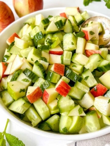 apple and cucumber salad in a white bowl with a spoon in it next to red apples and mint leaves.