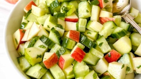 apple and cucumber salad in a white bowl with a spoon in it next to red apples and mint leaves.