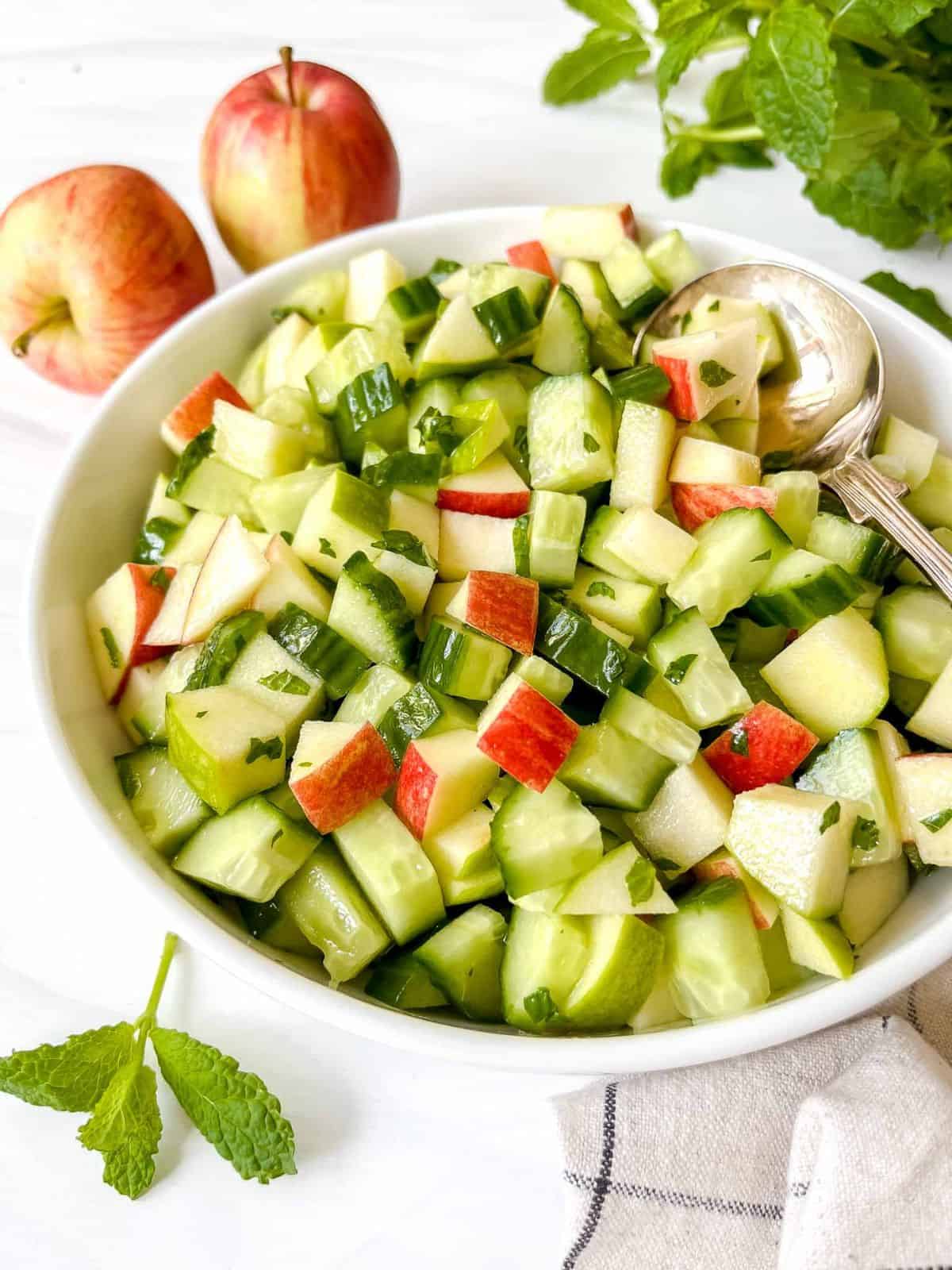 apple and cucumber salad in a white bowl next to mint leaves and red apples.