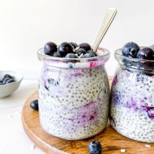 blueberry coconut chia pudding in two glass jars with spoons in it on a wooden board next to fresh blueberries.