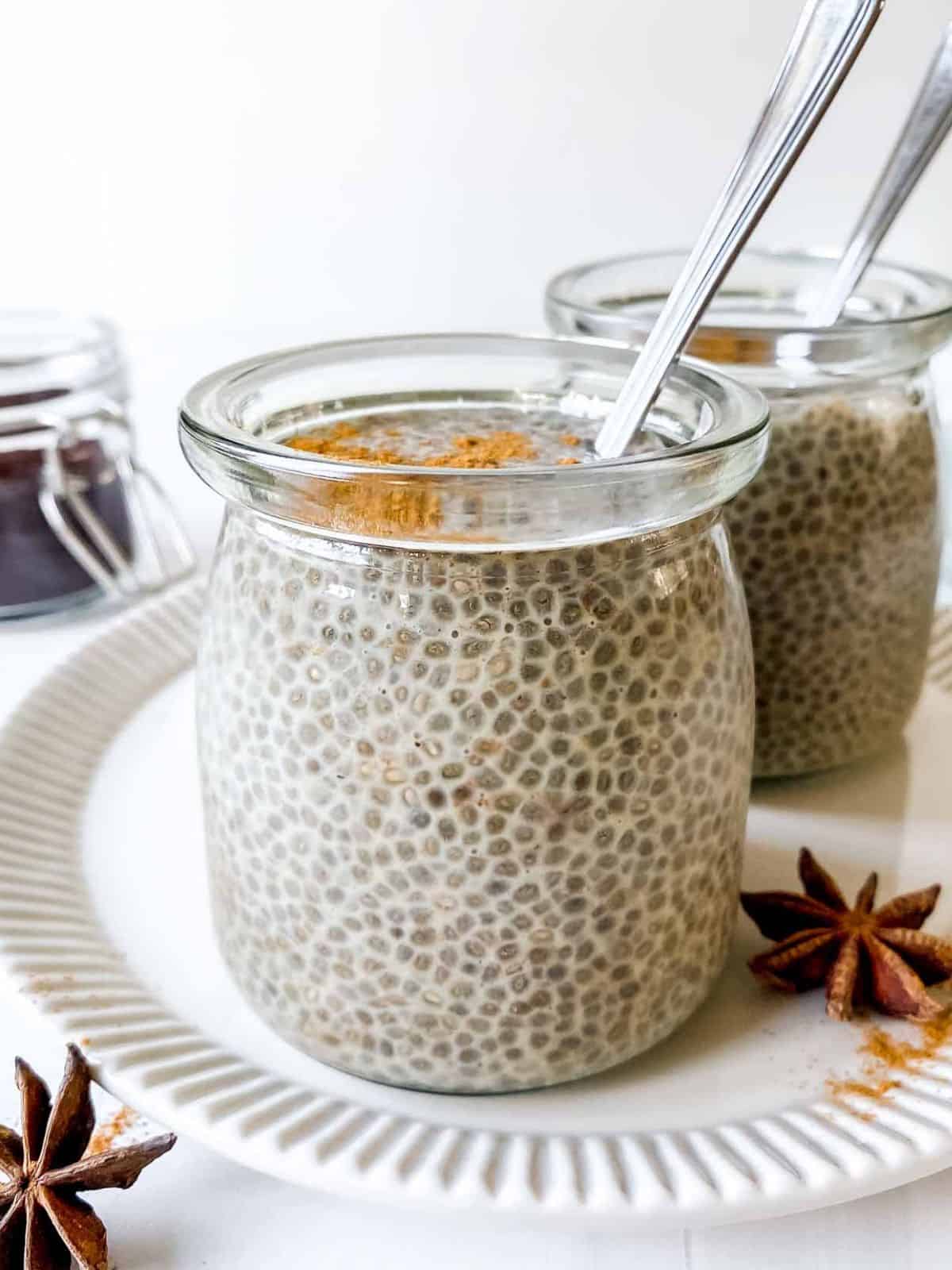 two glasses of coffee chia pudding with spoons in them on a cream plate with a jar of coffee in the background.