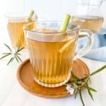 lemongrass ginger tea in two glasses on a round wooden plate with a blue cloth and jug in the background.