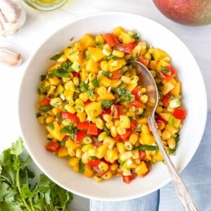 mango corn salsa in a white bowl with a spoon in it next to a blue cloth, cilantro and garlic.