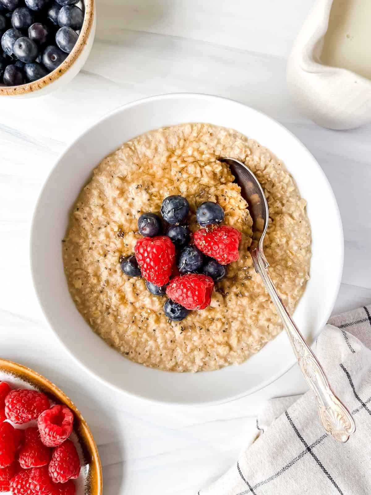 oat milk porridge in a white bowl with a spoon in it next to a bowl of blueberries and raspberries and a jug.