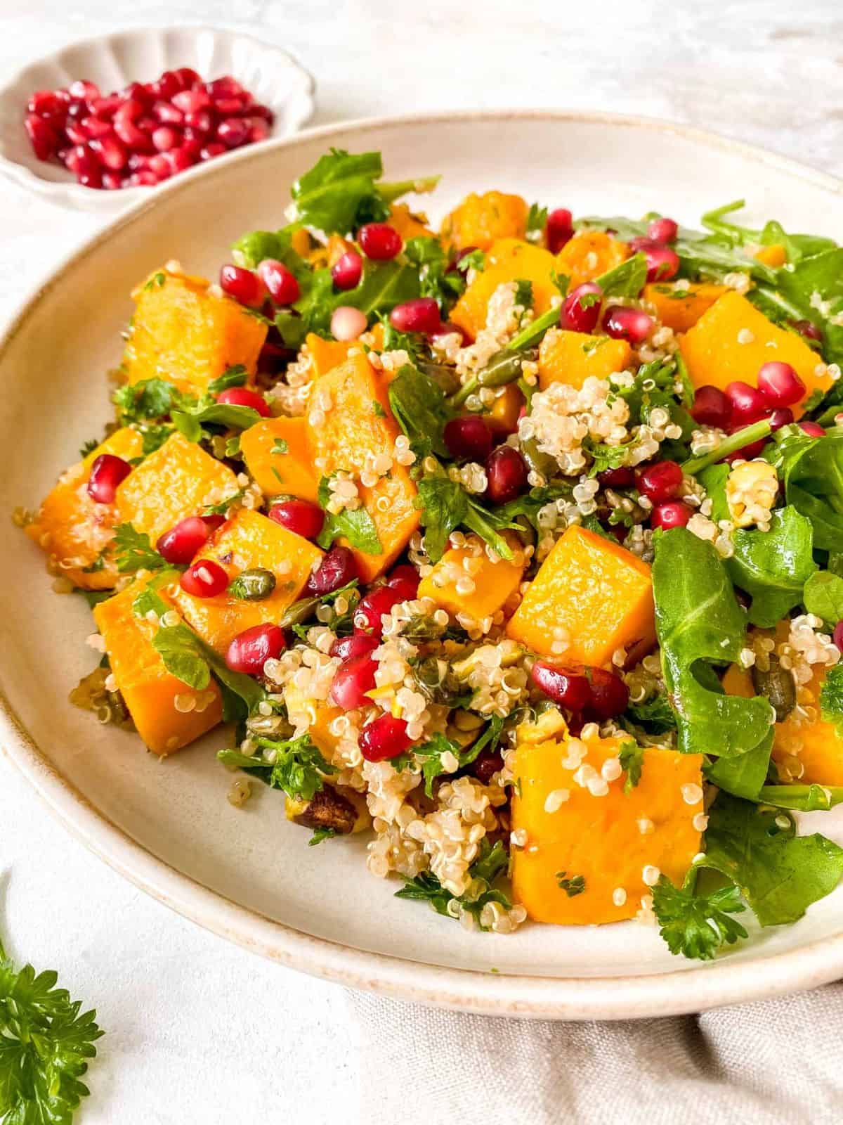 pumpkin quinoa salad on a light cream plate with a plate of pomegranate seeds in the background.