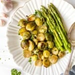 roasted asparagus and Brussels sprouts on a white plate with a spoon next to garlic cloves.