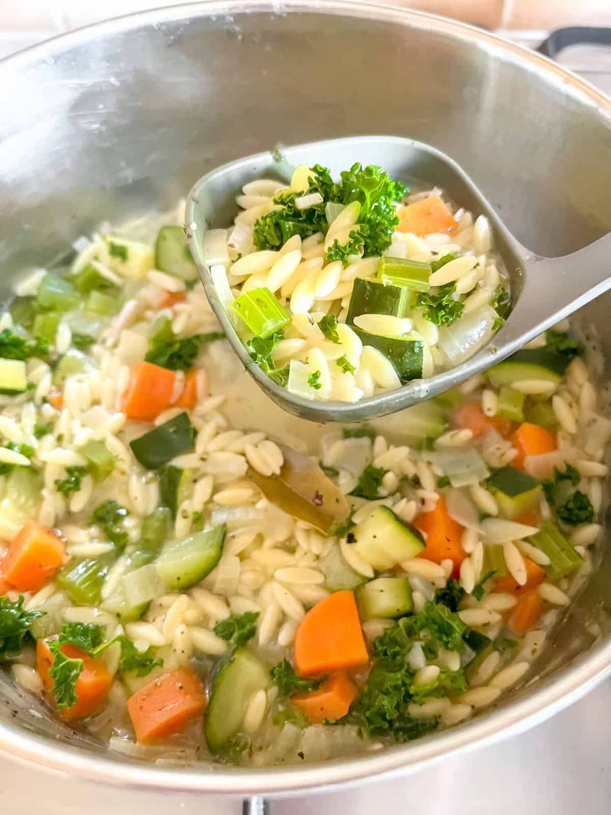 vegetable orzo soup in a ladle over a soup pot.