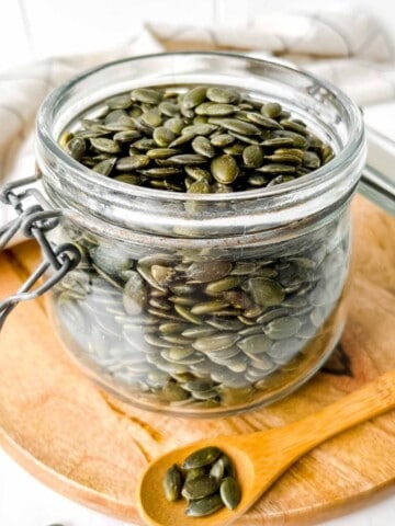 glass jar of pumpkin seeds on a wooden board next to a small wooden spoon of seeds.