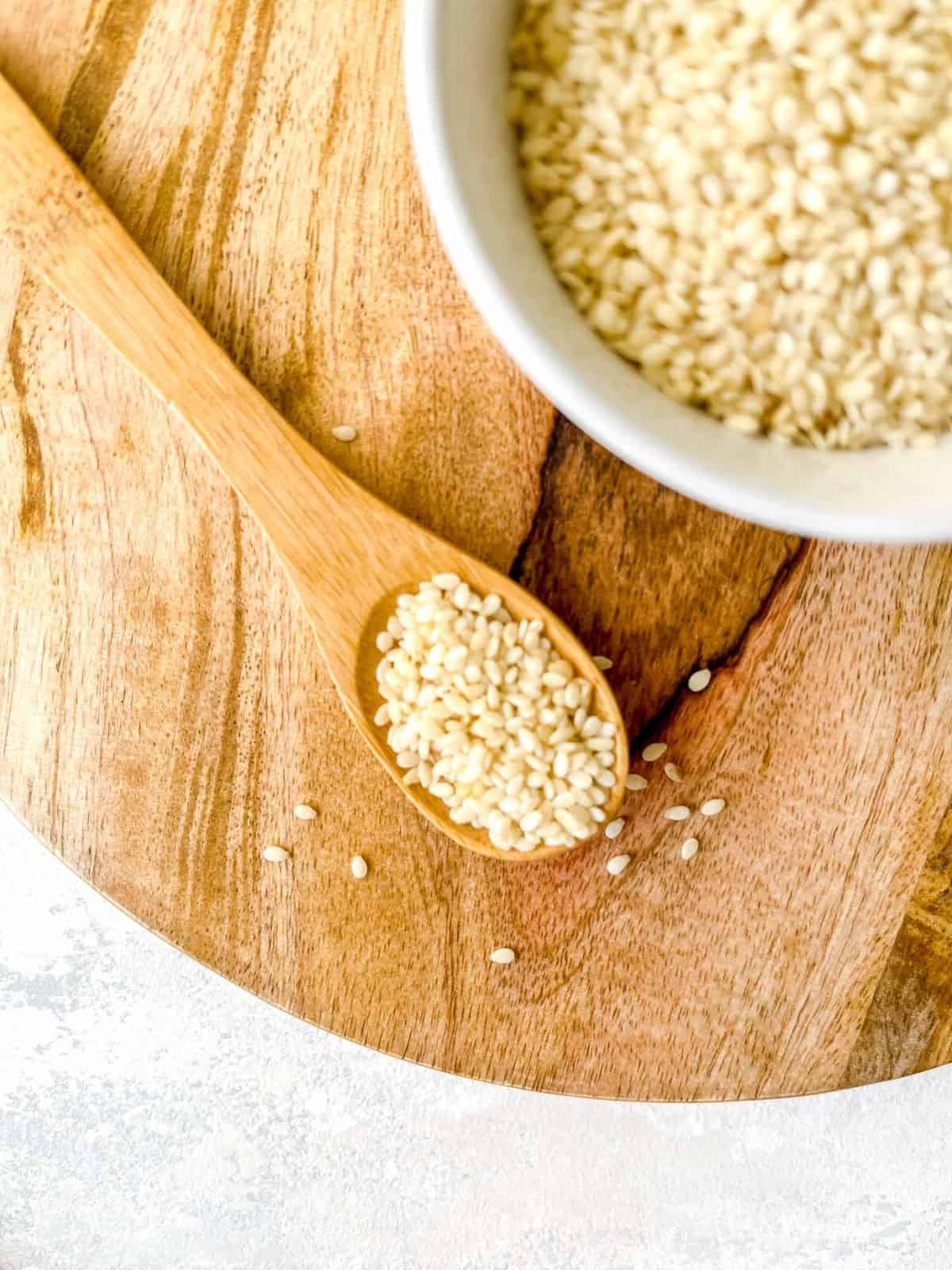 sesame seeds in a white bowl and on a wooden spoon on a wooden board.