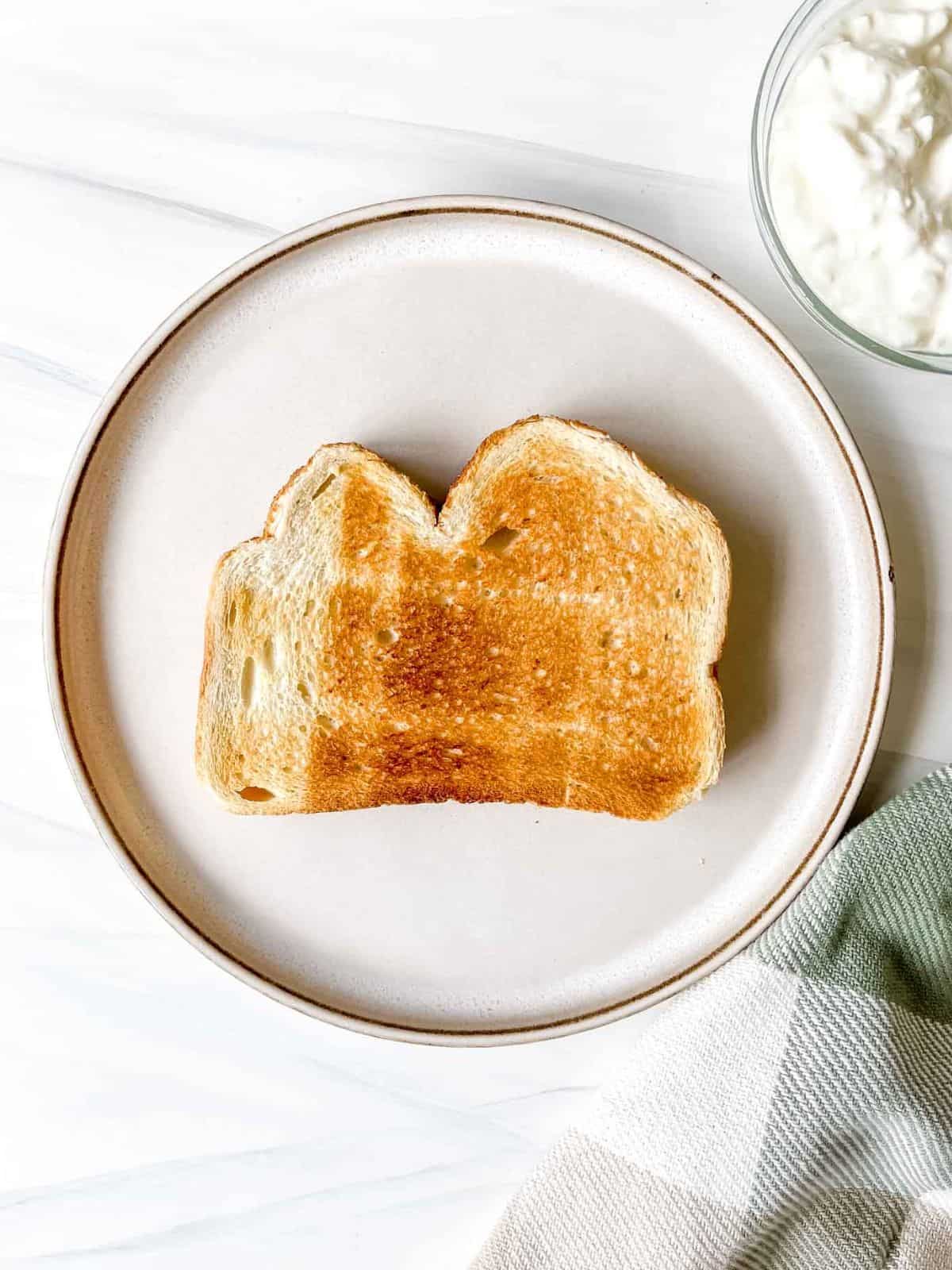 slice of toasted bread on a cream plate next to a bowl of cottage cheese and a green and white checked cloth.