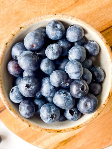 blueberries in a cream bowl on a wooden board next to blueberries.