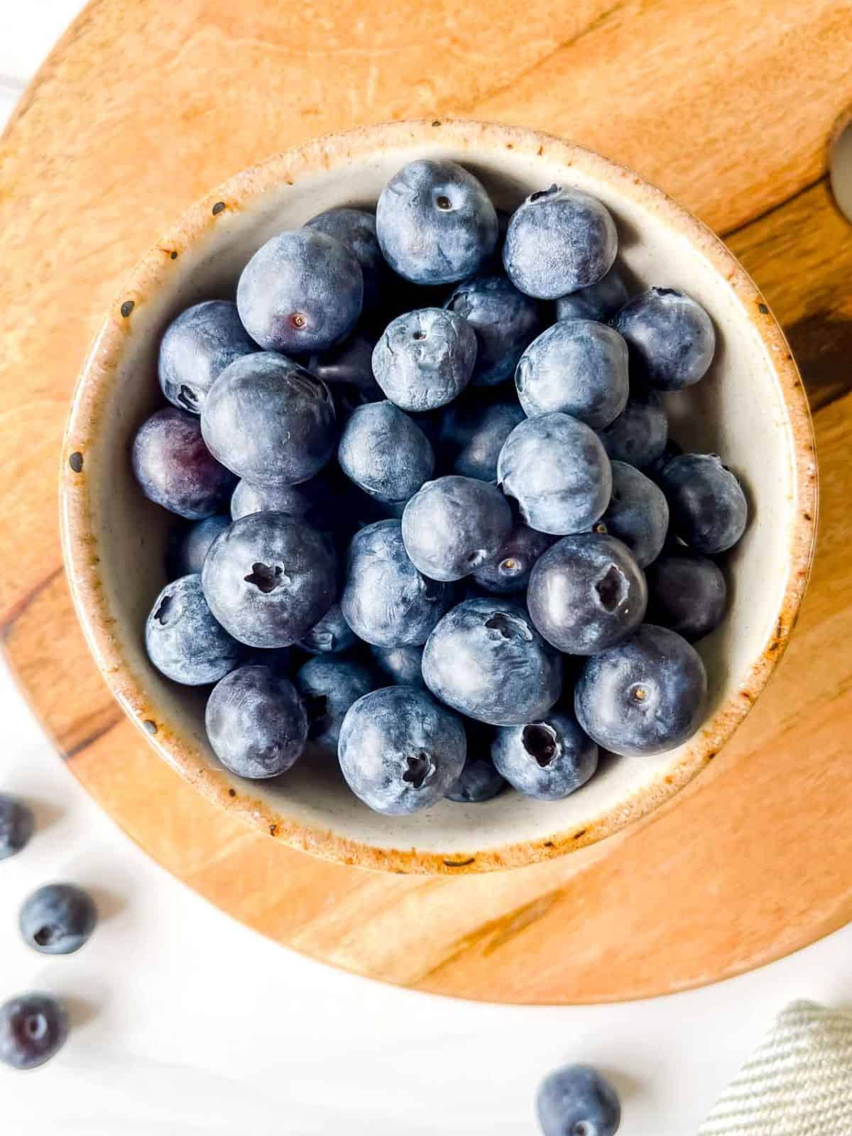 blueberries in a light cream bowl on a wooden board next to blueberries.